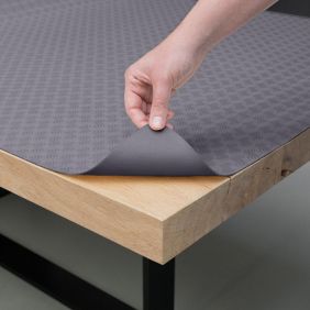 Een hand tilt een grijze PVC tafelbeschermer met een geometrisch patroon op van een houten tafelblad om de onderkant van het materiaal te tonen.